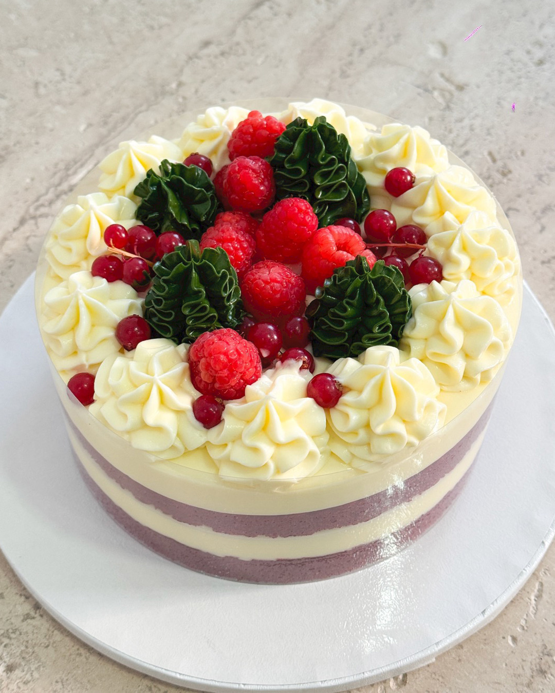 A round Red Velvet Christmas Cake with white and purple stripes, topped with swirls of white cream, fresh raspberries, redcurrants, and green decorative icing, displayed on a white board.