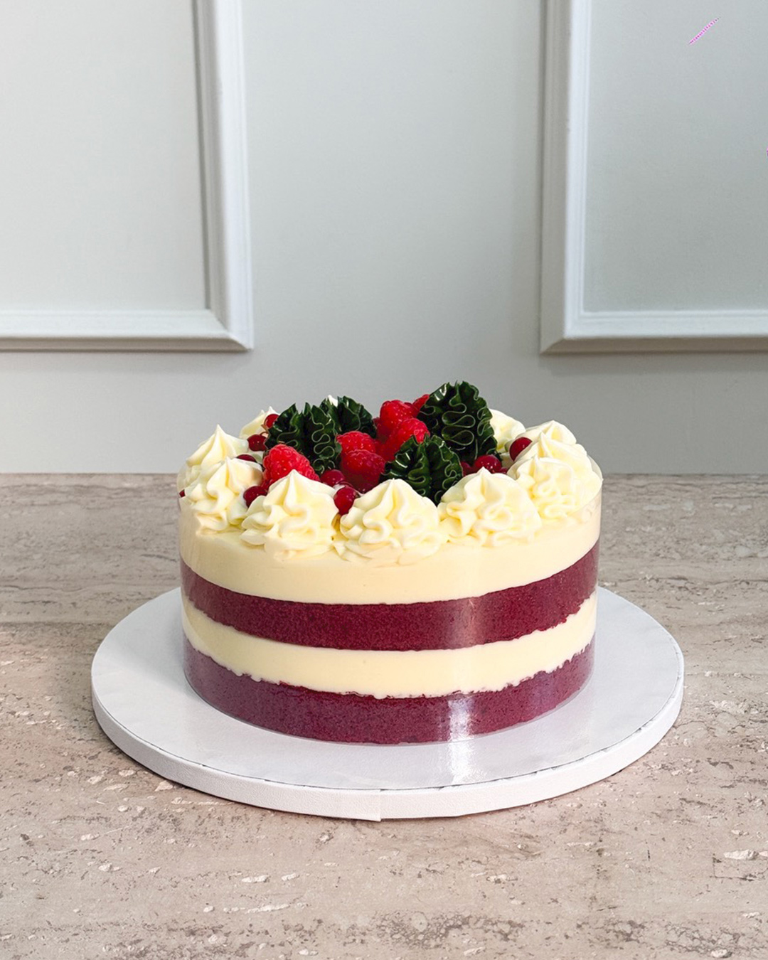 A round, layered Red Velvet Christmas Cake with white cream frosting, topped with whipped cream swirls, fresh raspberries, redcurrants, and green leaves, sits on a white cake board on a marble surface.