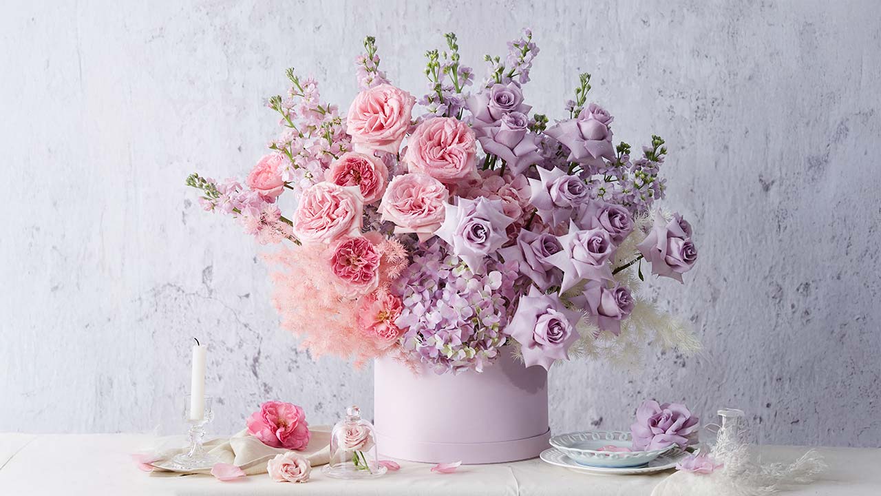 A round, pale pink box overflowing with pink and lavender roses, hydrangeas, and delicate fillers sits on a white table beside a candle, teacups, and scattered flower petals against a light grey background.