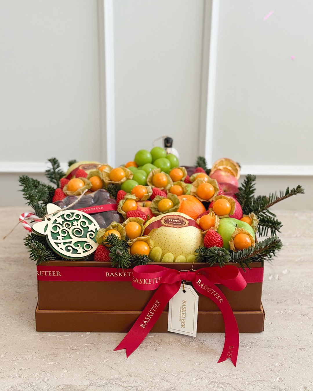 A festive fruit basket decorated with greenery, filled with assorted fresh fruits including grapes, apples, pears, and citrus fruits, and adorned with red ribbons and a decorative ornament, placed on a light-coloured surface.