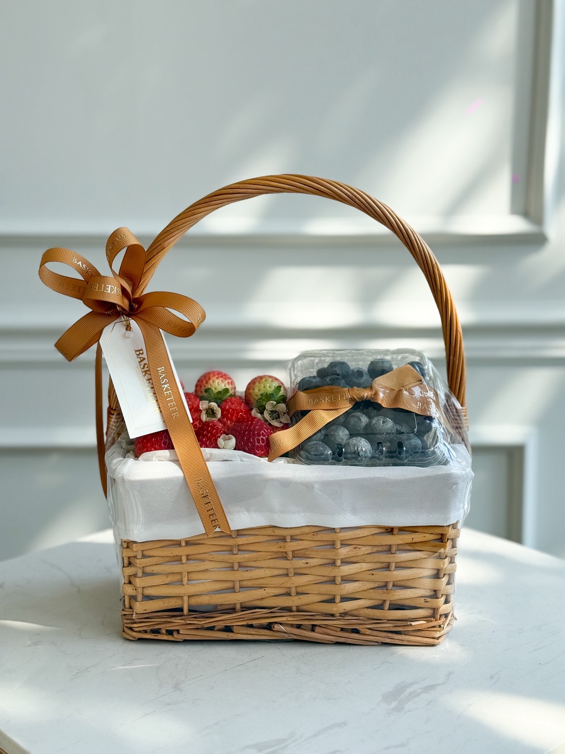 A Premium Strawberry & Blueberry Gift Hamper with a handle, lined with white cloth, holds fresh berries in clear containers. Adorned with a brown ribbon and a white tag, it’s perfect for gifting.