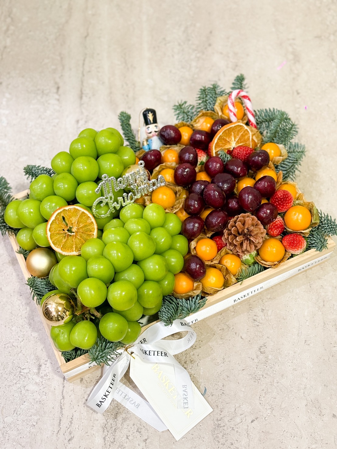 A wooden tray filled with green grapes, cherries, strawberries, gooseberries, dried orange slices, pine cones, and festive decorations, including pine sprigs and a “Merry Christmas” sign. White ribbon is tied to the tray.