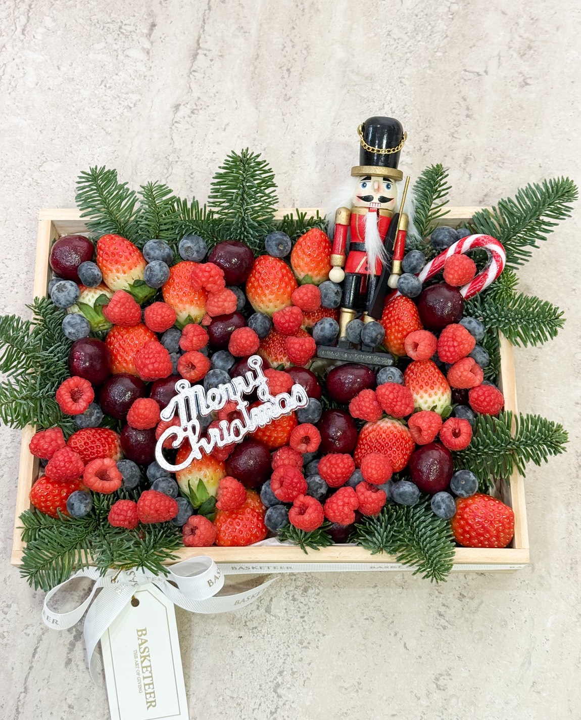 A wooden tray filled with assorted berries, cherries, and spruce branches, decorated with a 