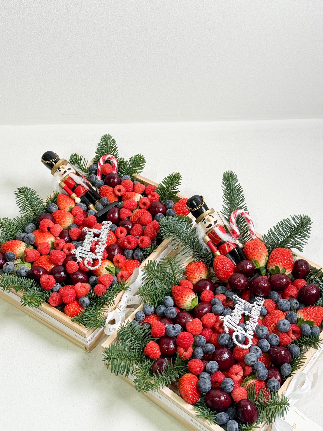 Two festive wooden trays filled with strawberries, raspberries, blueberries, and cherries, decorated with pine sprigs, candy canes, and nutcracker figurines, with a 