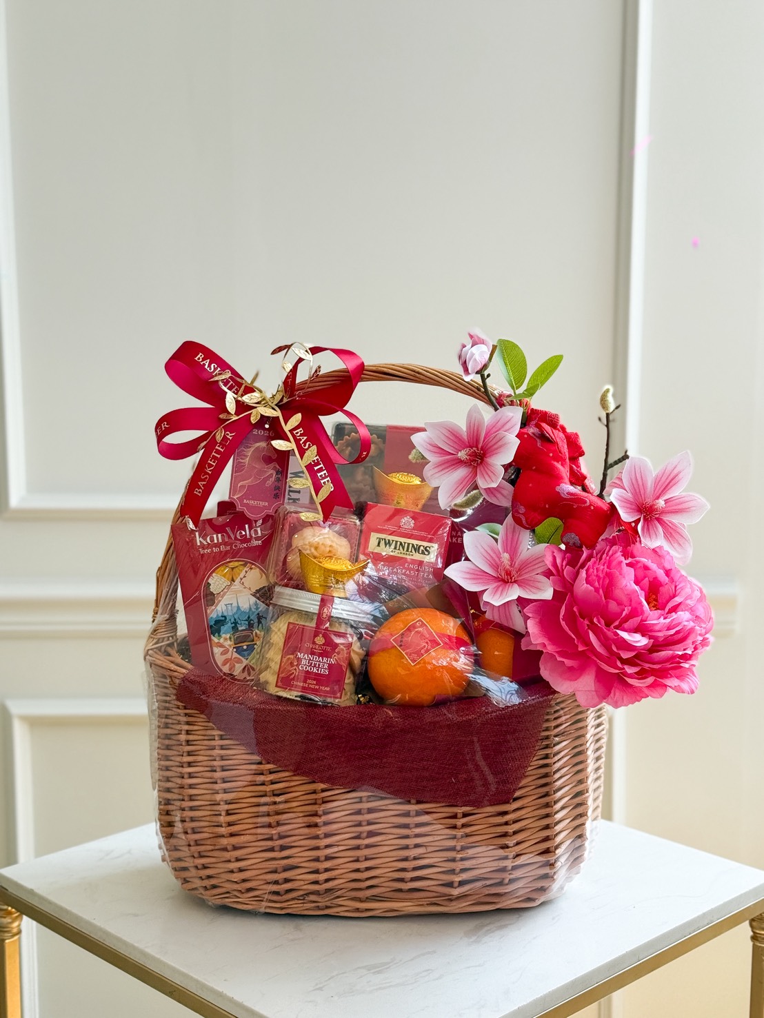 A wicker gift hamper wrapped in clear plastic, decorated with pink and red artificial flowers and a red ribbon, is filled with Mandarin Oranges with Premium Snacks, assorted tea, and fruit, displayed on a small white table.