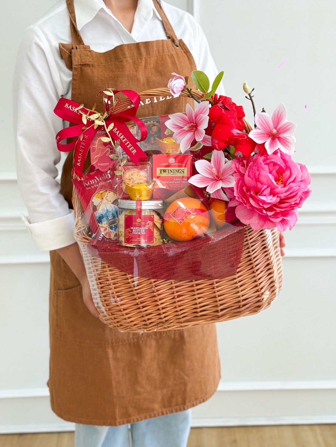 A person in a brown apron holds a wicker gift hamper filled with mandarins with premium snacks, tea, pink artificial flowers, and festive decorations, wrapped in clear plastic and tied with a red ribbon.