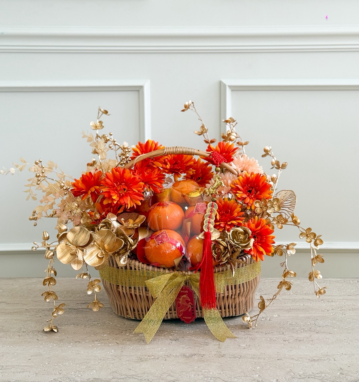 A decorative Chinese New Year Mandarin Orange Basket with golden floral accents, filled with red apples, orange flowers, and gold ornaments, adorned with a red tassel and gold ribbon on a light surface against a white panelled wall.