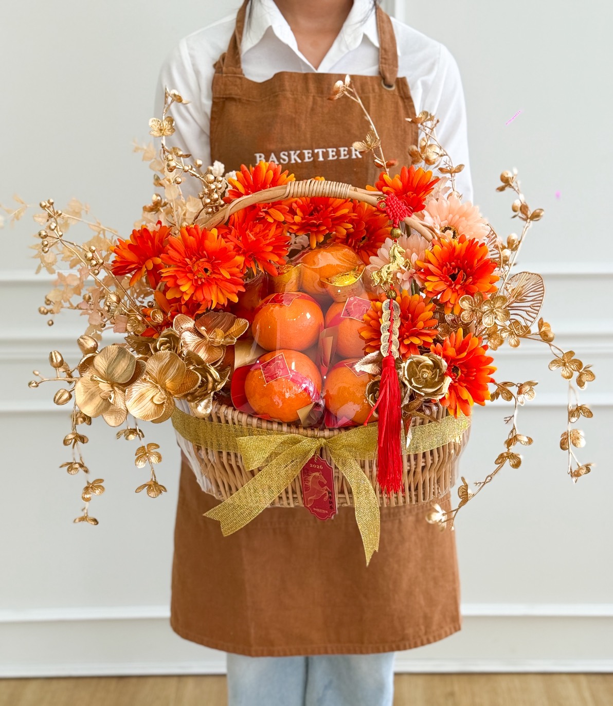 A person wearing a brown apron holds a Chinese New Year Mandarin Orange Basket with Golden Floral Accents, filled with bright orange flowers, decorative gold leaves, and oranges wrapped in netting, adorned with a gold ribbon and red tassel.