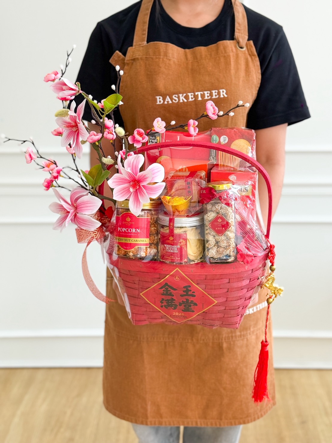 A person wearing a brown apron holds a Chinese New Year Gourmet Snacks Gift Hamper filled with snacks, dried goods, and pink artificial flowers. The festive hamper features Chinese characters and decorations, perfect for celebrating the season.