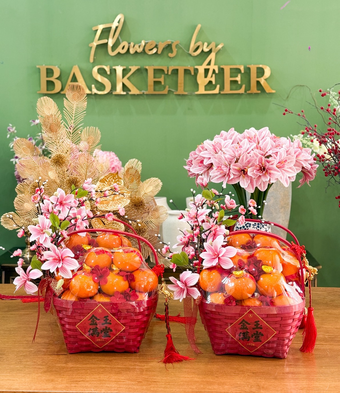 Two red Chinese New Year Gourmet Snacks Gift Hampers with artificial orange fruit and pink flowers are displayed on a wooden table. Behind them is a green wall with the gold sign 