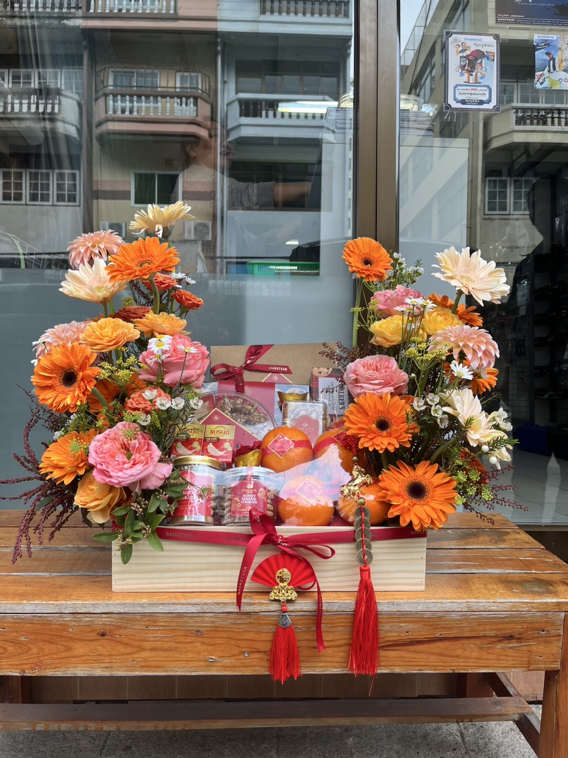 A Chinese New Year Luxury Gourmet & Wellness Hamper with vibrant orange and pink flowers, festive red ribbons, and gift items sits on a wooden table outside a glass building, with street views and flat balconies in the background.