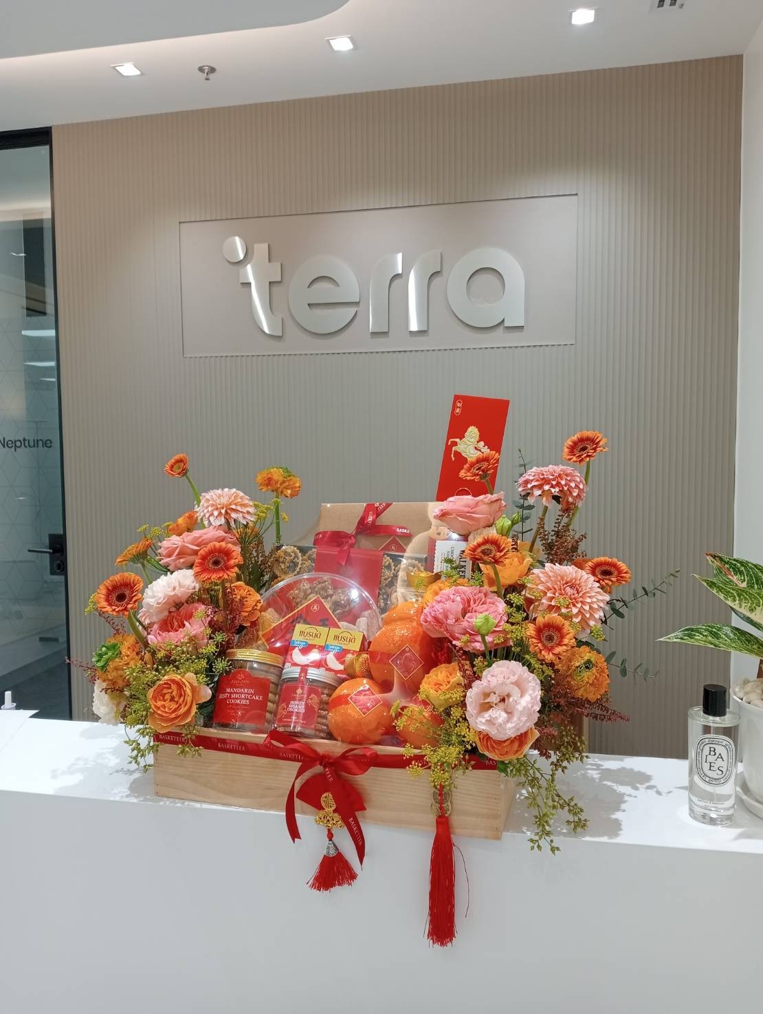 A Chinese New Year Luxury Gourmet & Wellness Gift Hamper brimming with colourful flowers, festive packaging, and red envelopes sits elegantly on a white counter in front of the “terra” logo, creating a celebratory atmosphere.