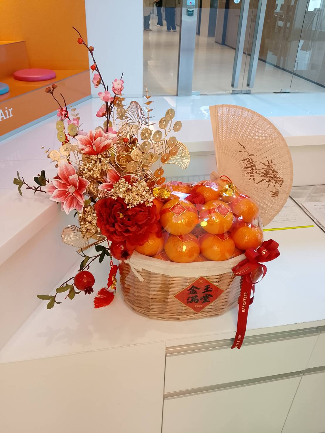 A Chinese New Year Mandarin Orange Basket with Bamboo Fan, adorned with red and pink artificial flowers, gold accents, and festive red ribbons—perfect for celebrating the season in vibrant style.