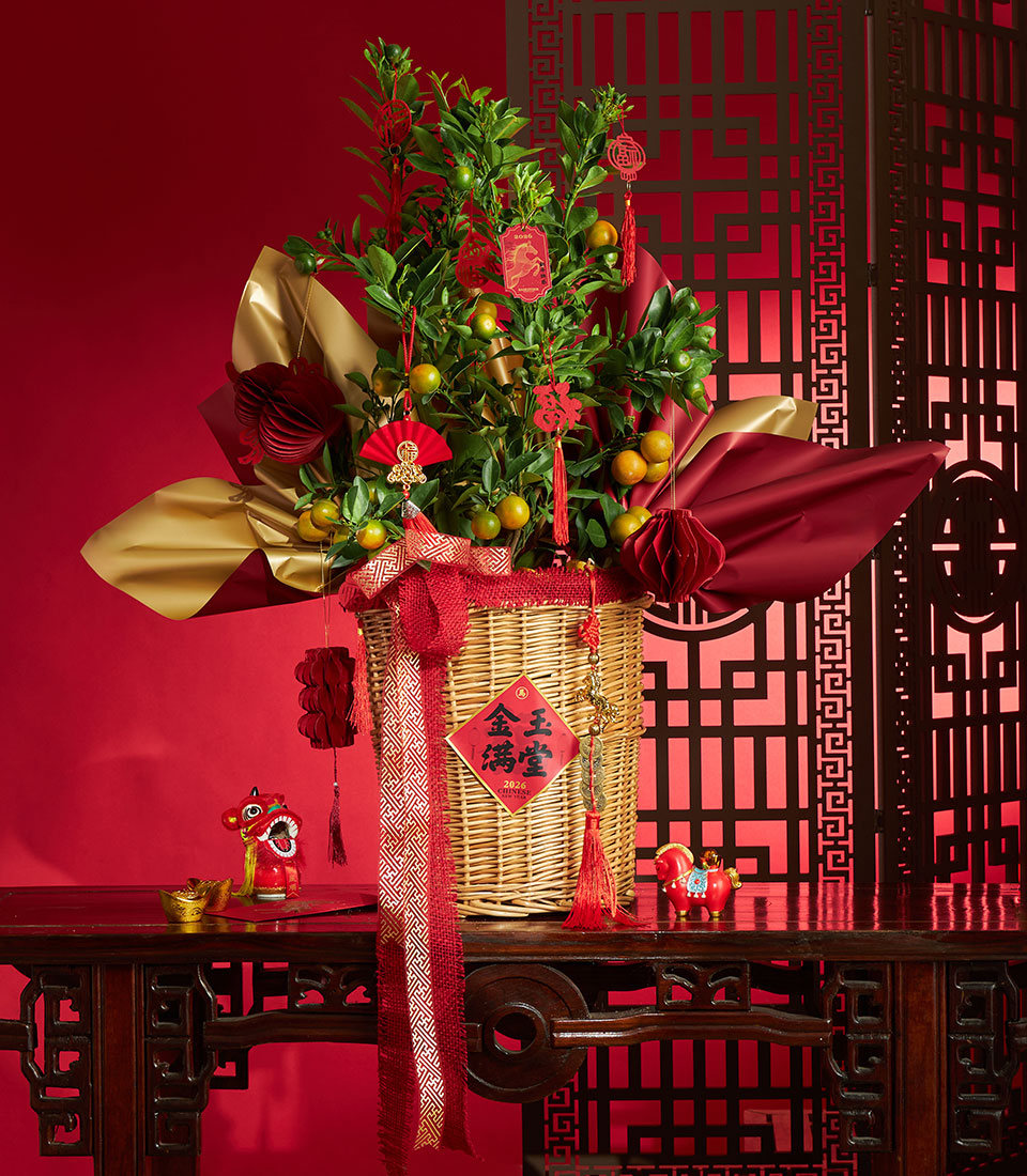 A festive Chinese New Year arrangement with a wicker basket of mandarin plants, red and gold decorations, and lucky charms on a wooden table, set against a red background with traditional Chinese patterns.