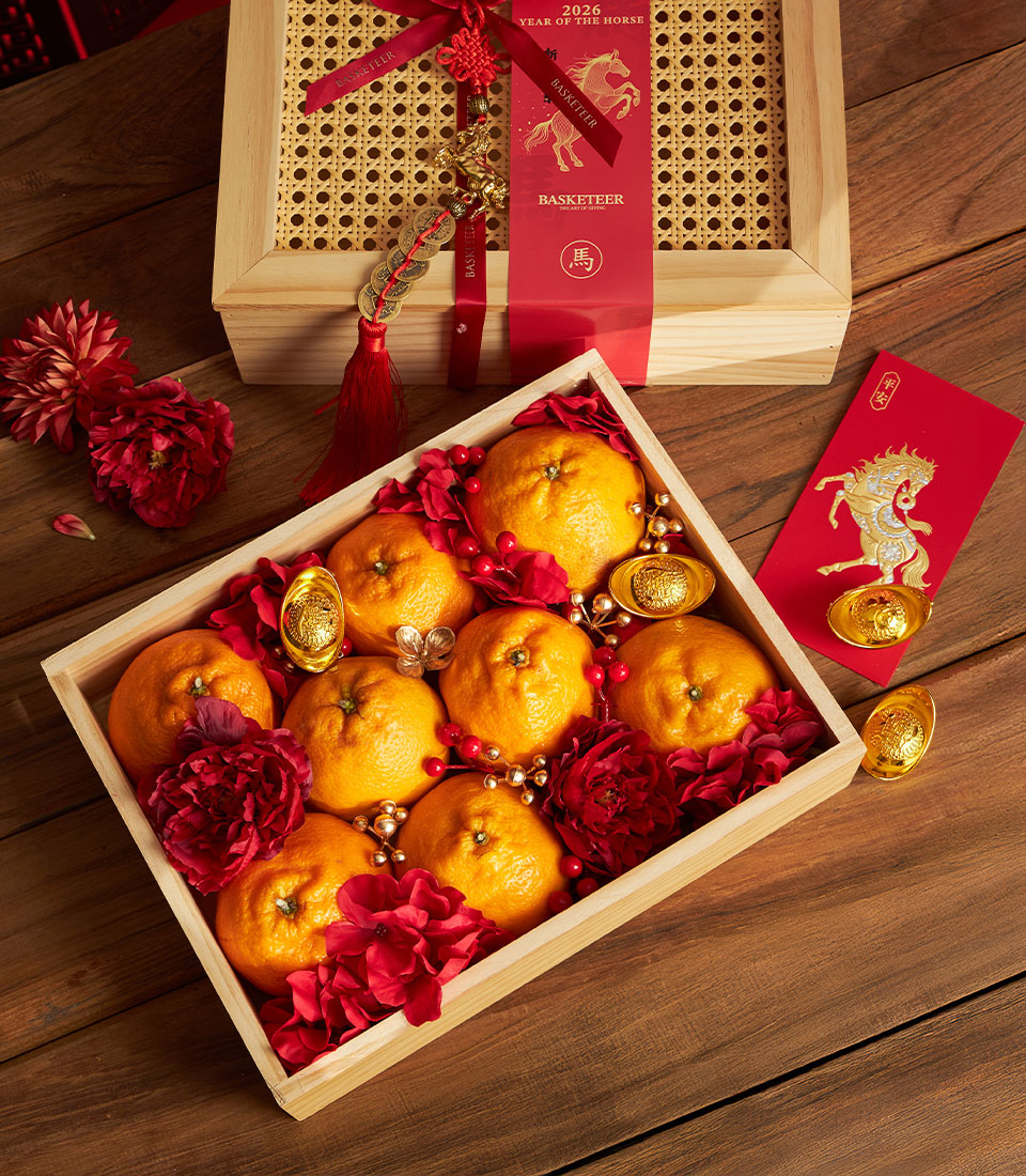 A Dekopon Orange Prosperity Gift Box filled with oranges, red flowers, gold ingots, and beads sits on a wooden table beside a red envelope and a decorated gift box for Lunar New Year.