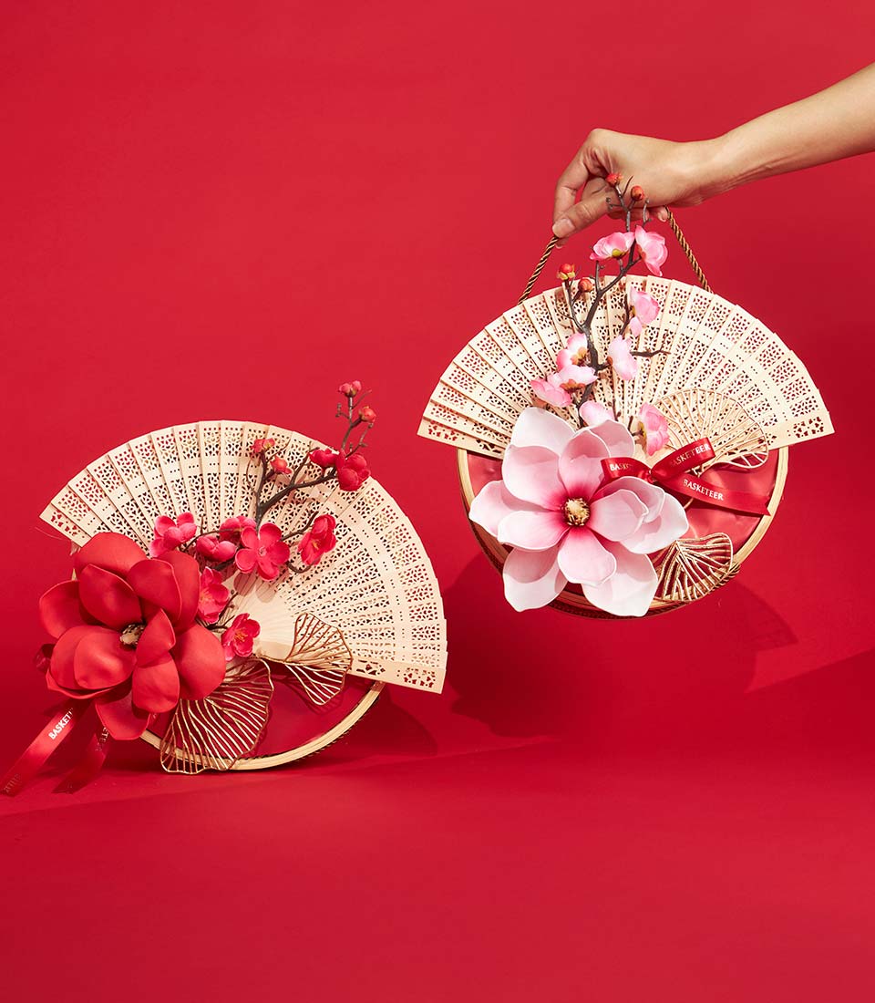 Two decorative hand fans with intricate patterns, adorned with large artificial red and pink flowers, are displayed against a red background. A hand holds one fan, whilst the other rests on the surface.