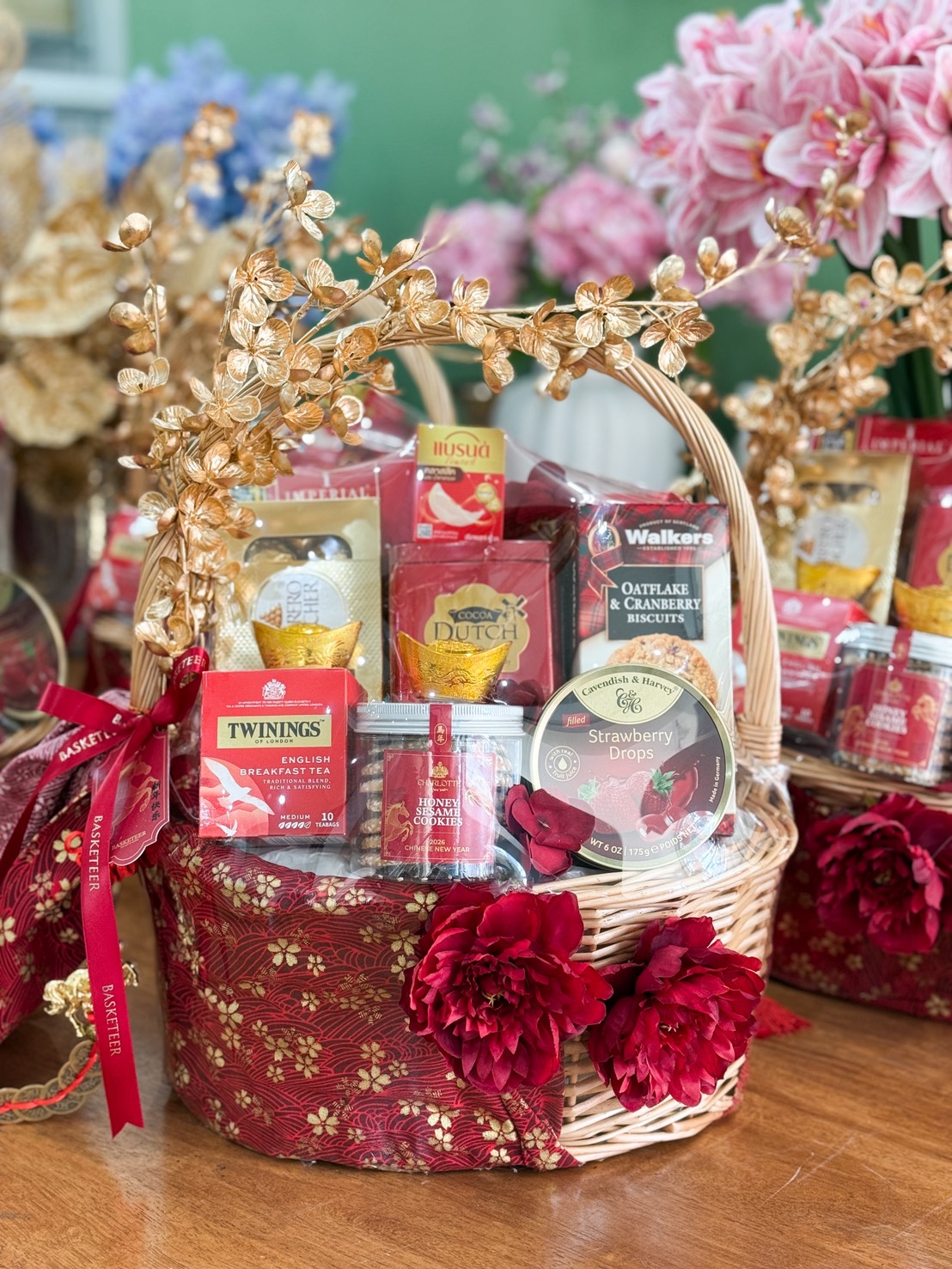 The Golden Fortune Gourmet Gift Hamper is a decorative gift hamper filled with assorted snacks, tea, and sweets, wrapped in red fabric with gold floral accents, red flowers, and a gold floral ornament, set on a wooden table with pink flowers in the background.