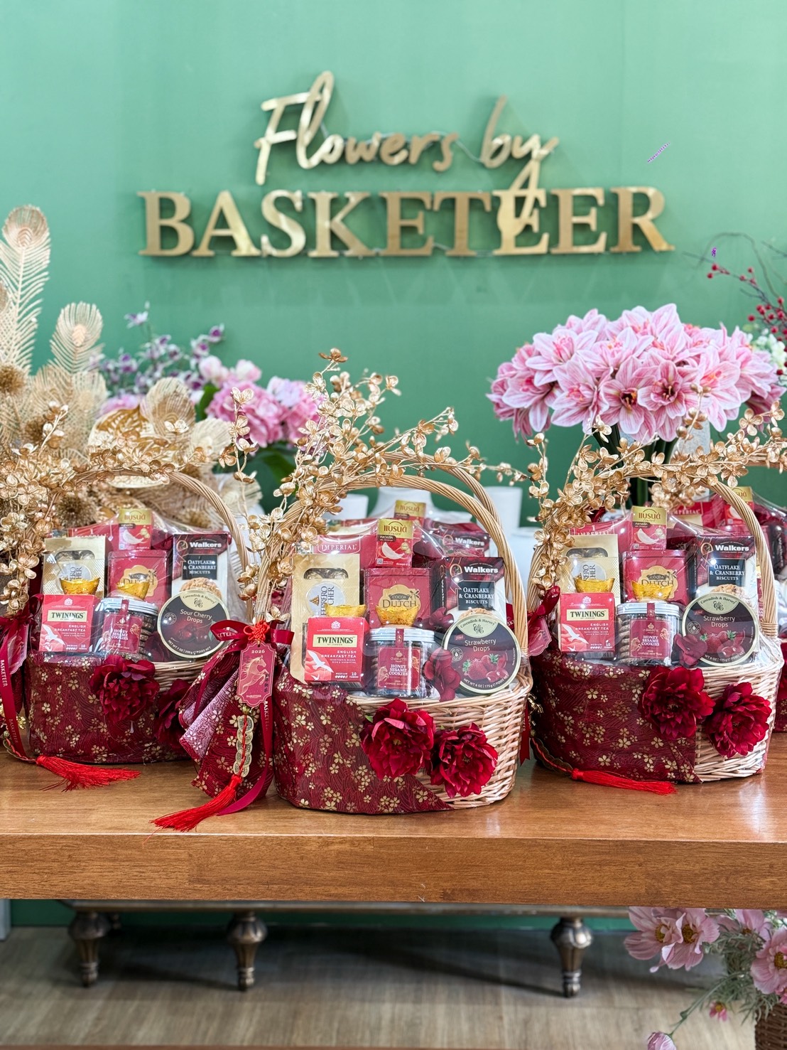 Three decorative Golden Fortune Gourmet Gift Hampers filled with assorted snacks and drinks are displayed on a wooden table. Each hamper is adorned with red fabric, gold accents, and artificial flowers. A green wall behind reads “Flowers by Basketeer.”.