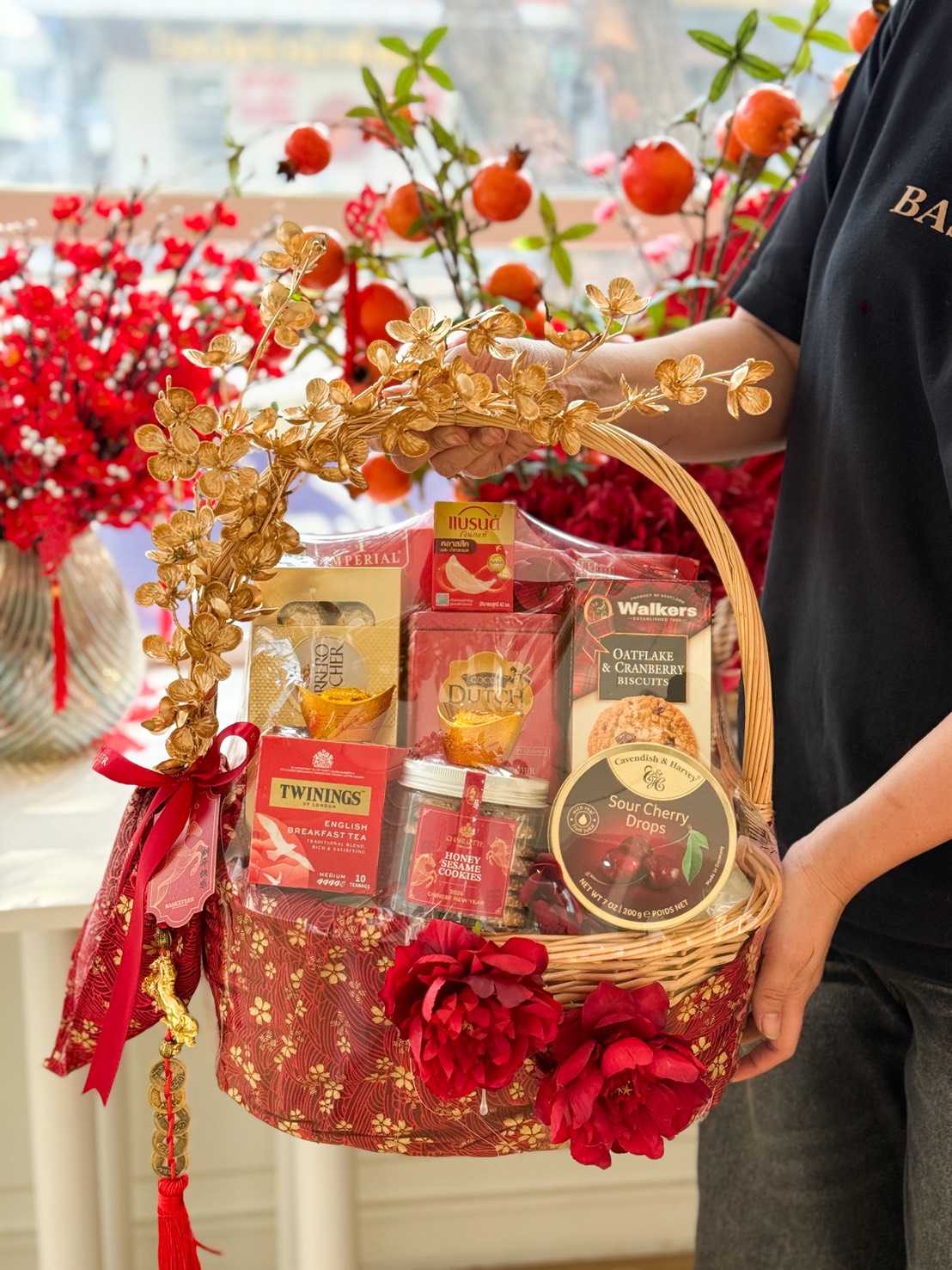 A person holds a Golden Fortune Gourmet Gift Hamper adorned with red and gold flowers, filled with assorted snacks, tea, and biscuits. Red blossoms and fruit branches in the background create a vibrant, celebratory display.