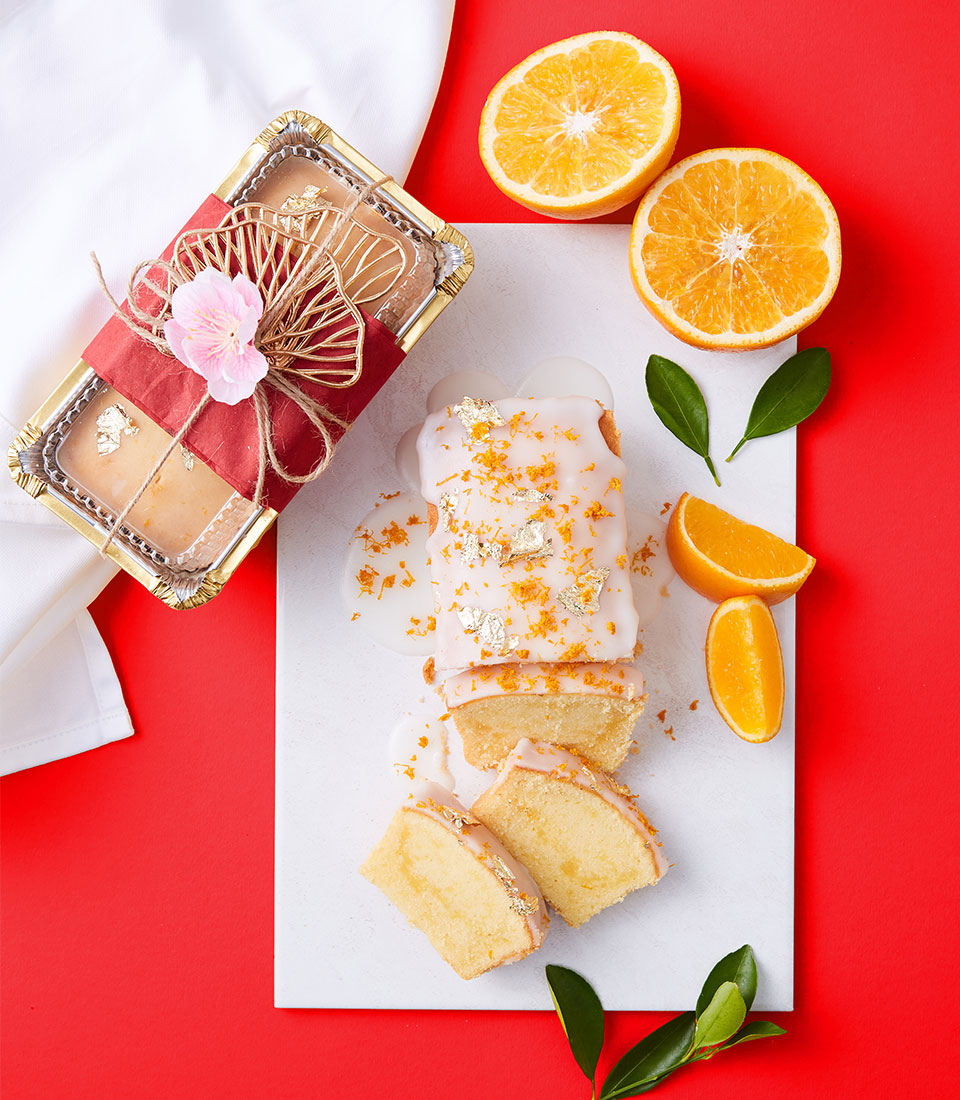 A loaf of glazed orange pound cake, partially sliced, is displayed on a white board with orange slices and green leaves. A whole orange, halved, and a decorated loaf wrapped with red ribbon sit nearby on a red background.