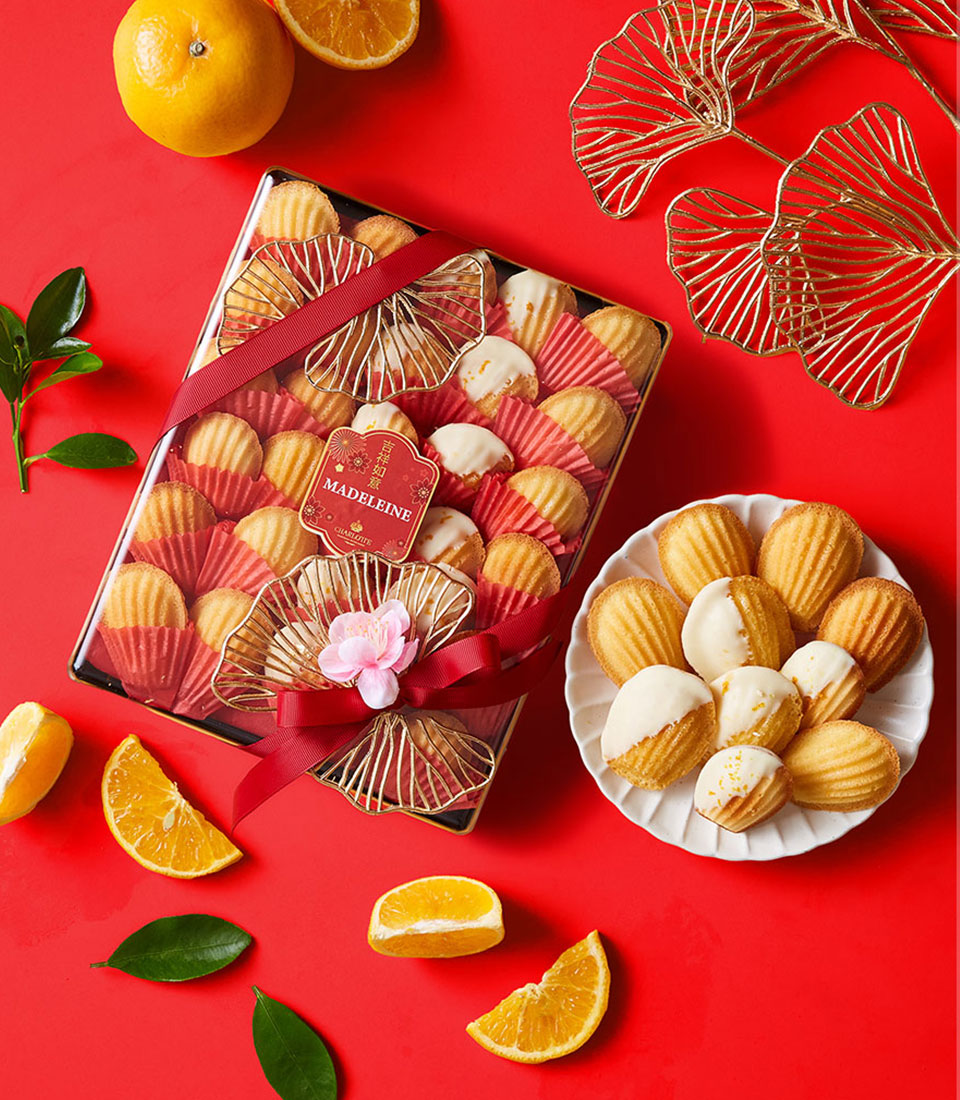 A box of assorted madeleines tied with a red ribbon, a white plate of madeleines, orange slices, whole oranges, green leaves, and gold ginkgo leaf decorations on a red background.