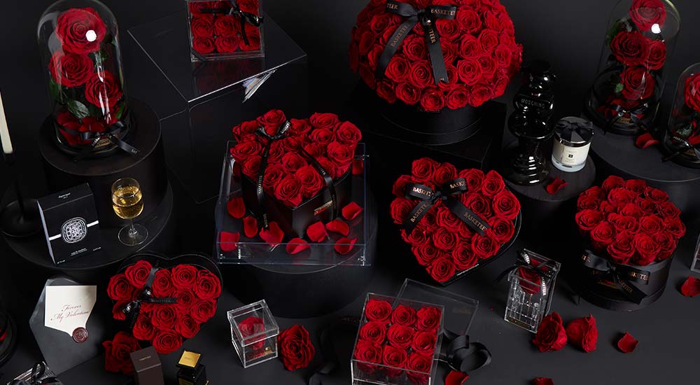 A luxurious arrangement of red roses in various shapes—boxes, heart, domes—displayed with black gift boxes, glass containers, candles, and scattered rose petals on a dark surface.