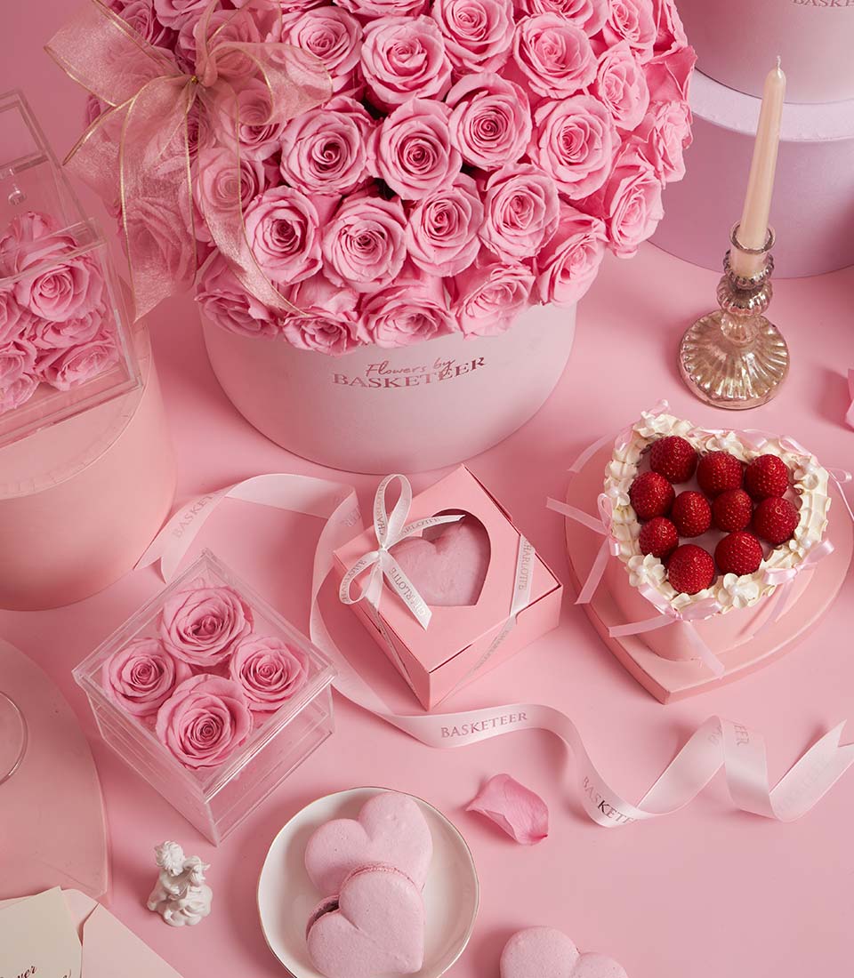 A pastel pink scene perfect for a homepage, featuring a large bouquet of pink roses, heart-shaped raspberry cake, pink heart biscuits, a candle, small rose arrangements, and decorative ribbon and petals—ideal for select popular occasions.