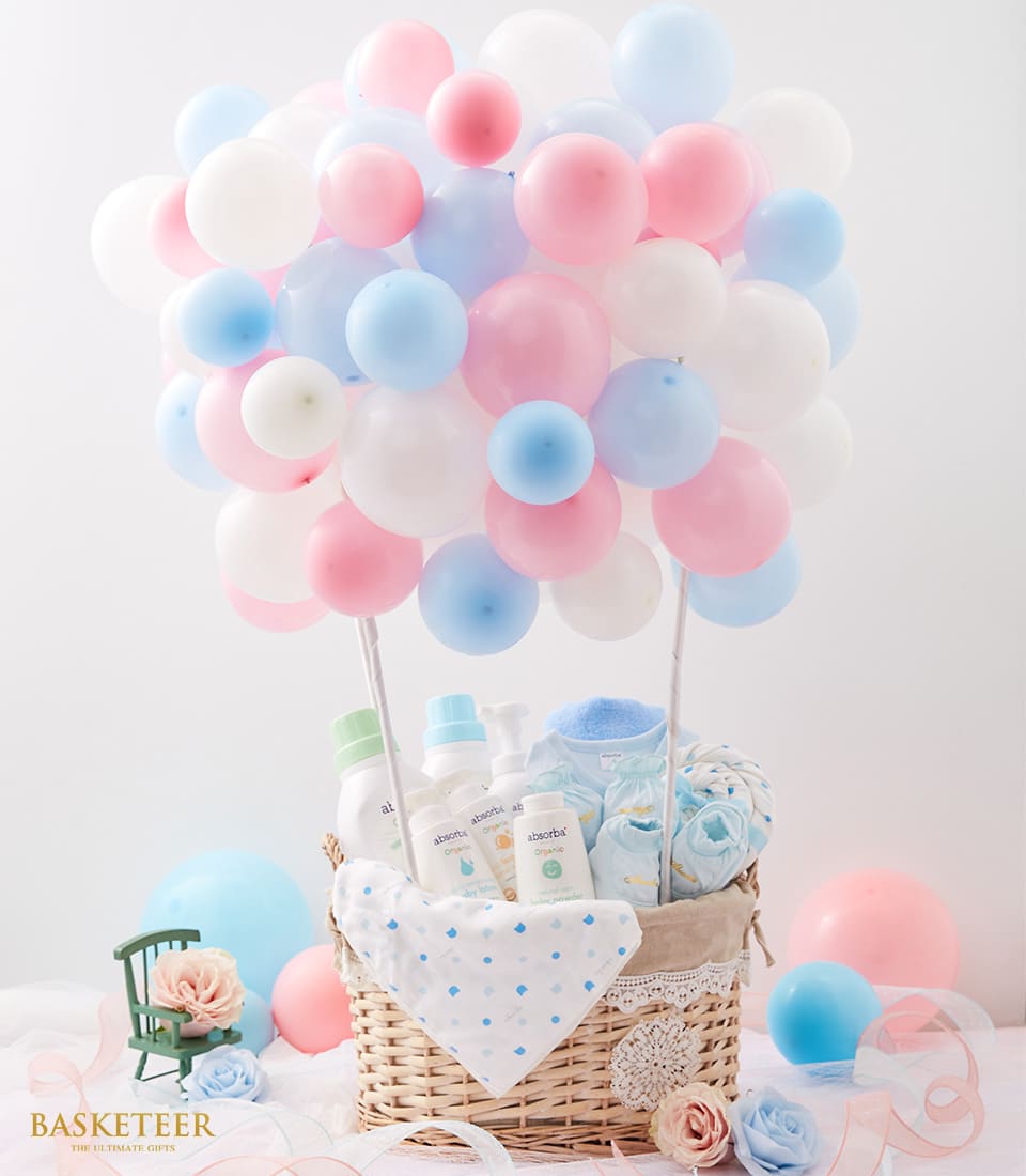 A wicker basket filled with baby care products, towels, and blankets, topped with pink, blue, and white balloons. Pastel balloons surround the basket. The Basketeer logo is visible in the bottom corner.