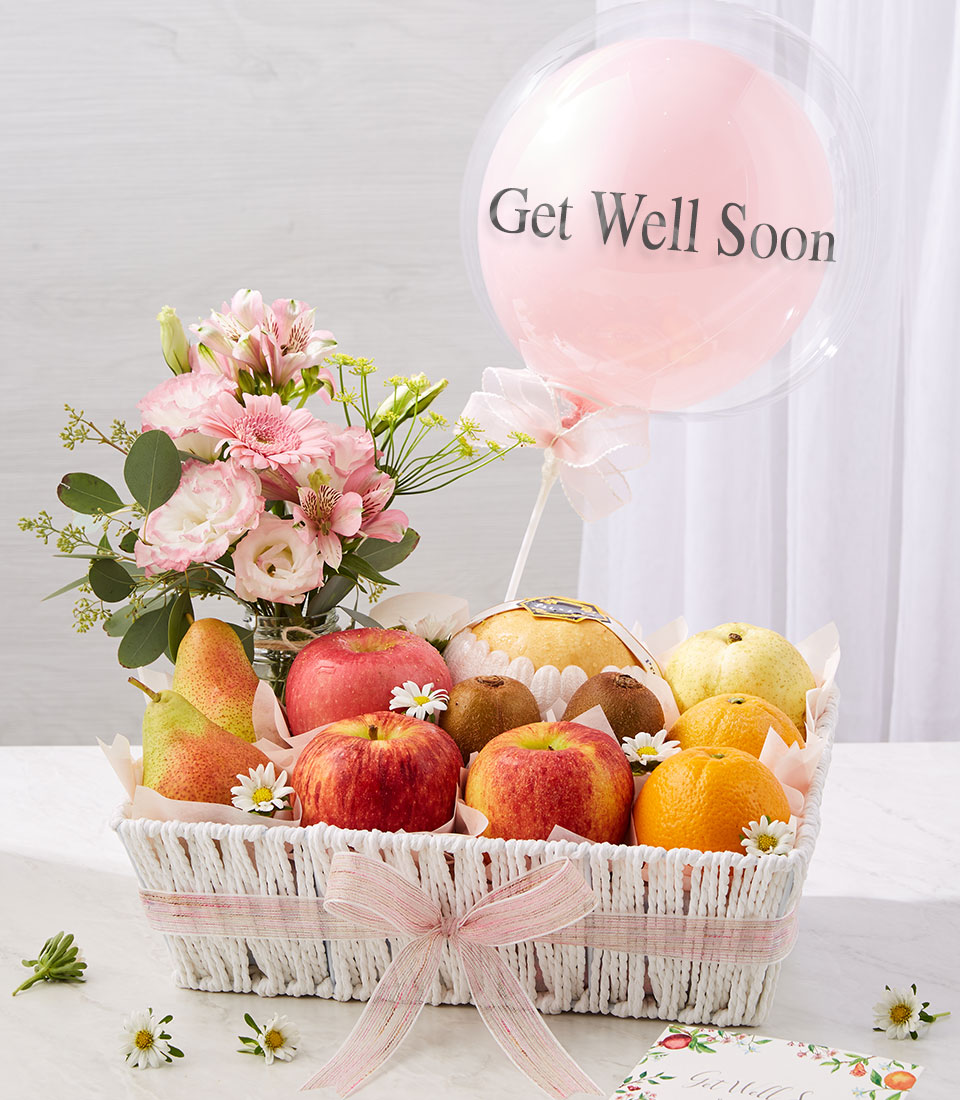 A white basket with pink ribbon holds apples, oranges, pears, and small flowers. Next to it is a bouquet of pink flowers and a pink balloon with "Get Well Soon" written on it. A card is visible in the corner.