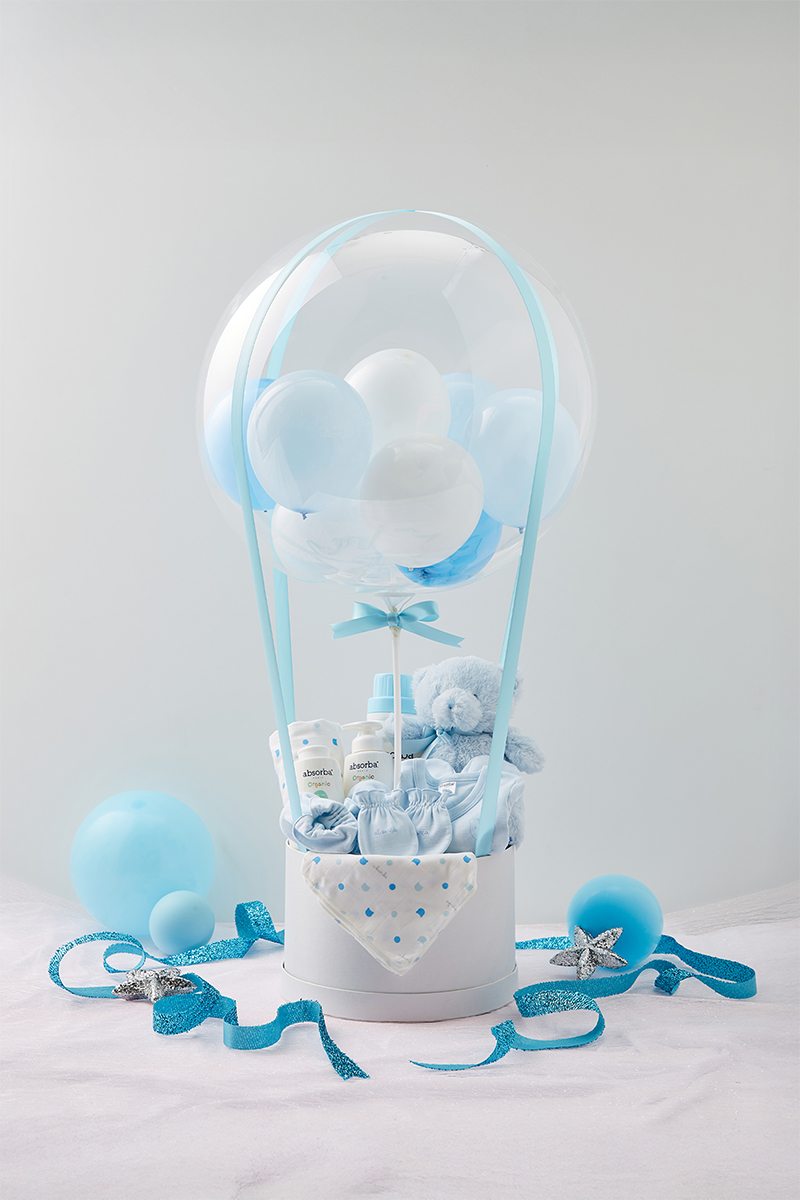 A white basket with baby items, blue and white balloons arranged like a hot air balloon, blue ribbons, soft toy, and baby care products, surrounded by blue balloons and silver star decorations on a light background.