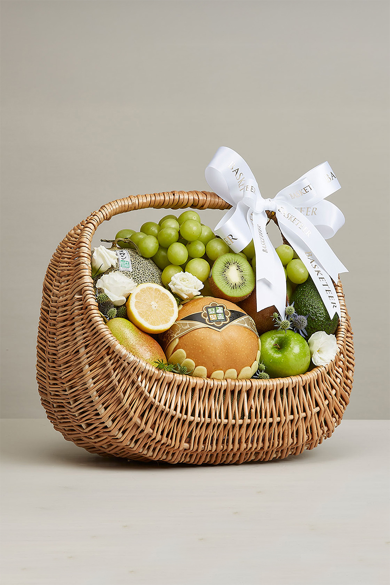 A wicker basket filled with green grapes, kiwi, green apple, lemon, pear, melon, and white flowers, adorned with a large white bow and ribbon, displayed on a light surface with a plain background.