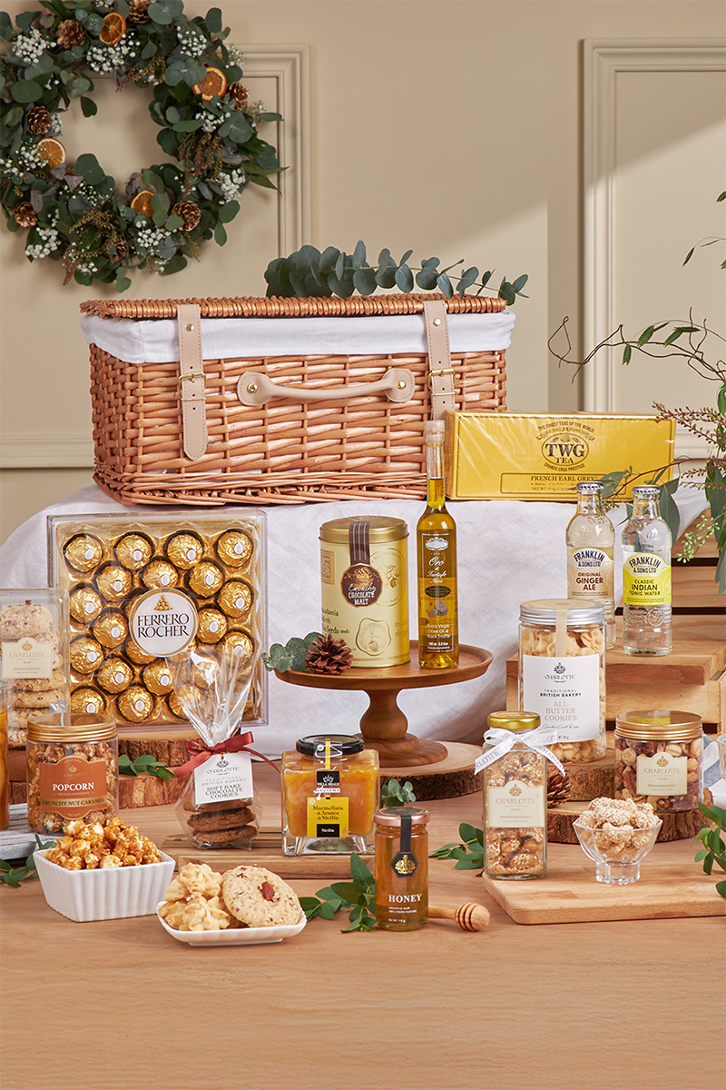 A wicker gift hamper sits on a table, surrounded by treats including Ferrero Rocher chocolates, honey, jam, popcorn, biscuits, breadsticks, olive oil, sparkling drinks, and loose leaf tea, with festive holiday decor in the background.