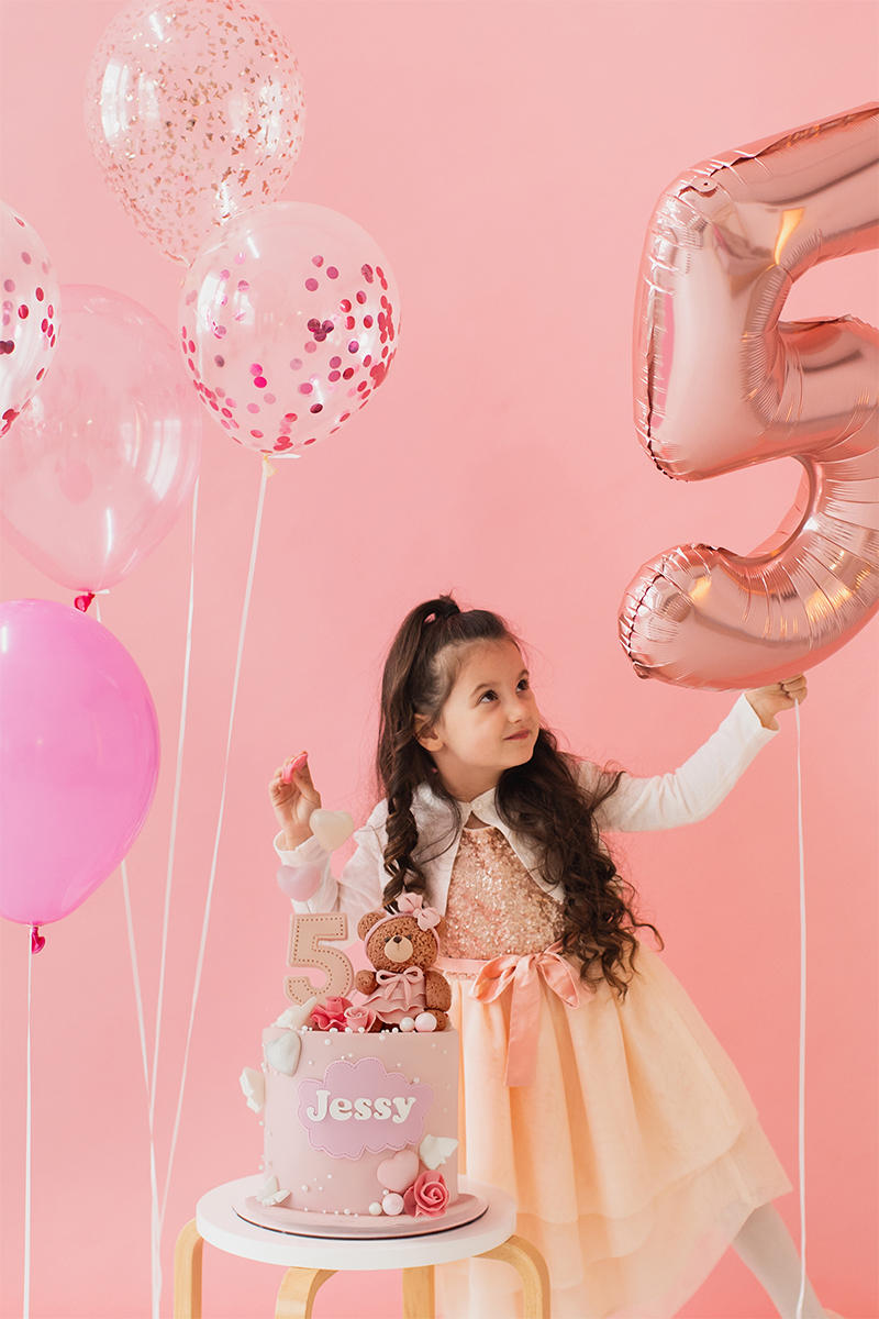 A young girl in a peach dress holds a large rose-gold balloon shaped like the number 5, standing next to a birthday cake labelled 