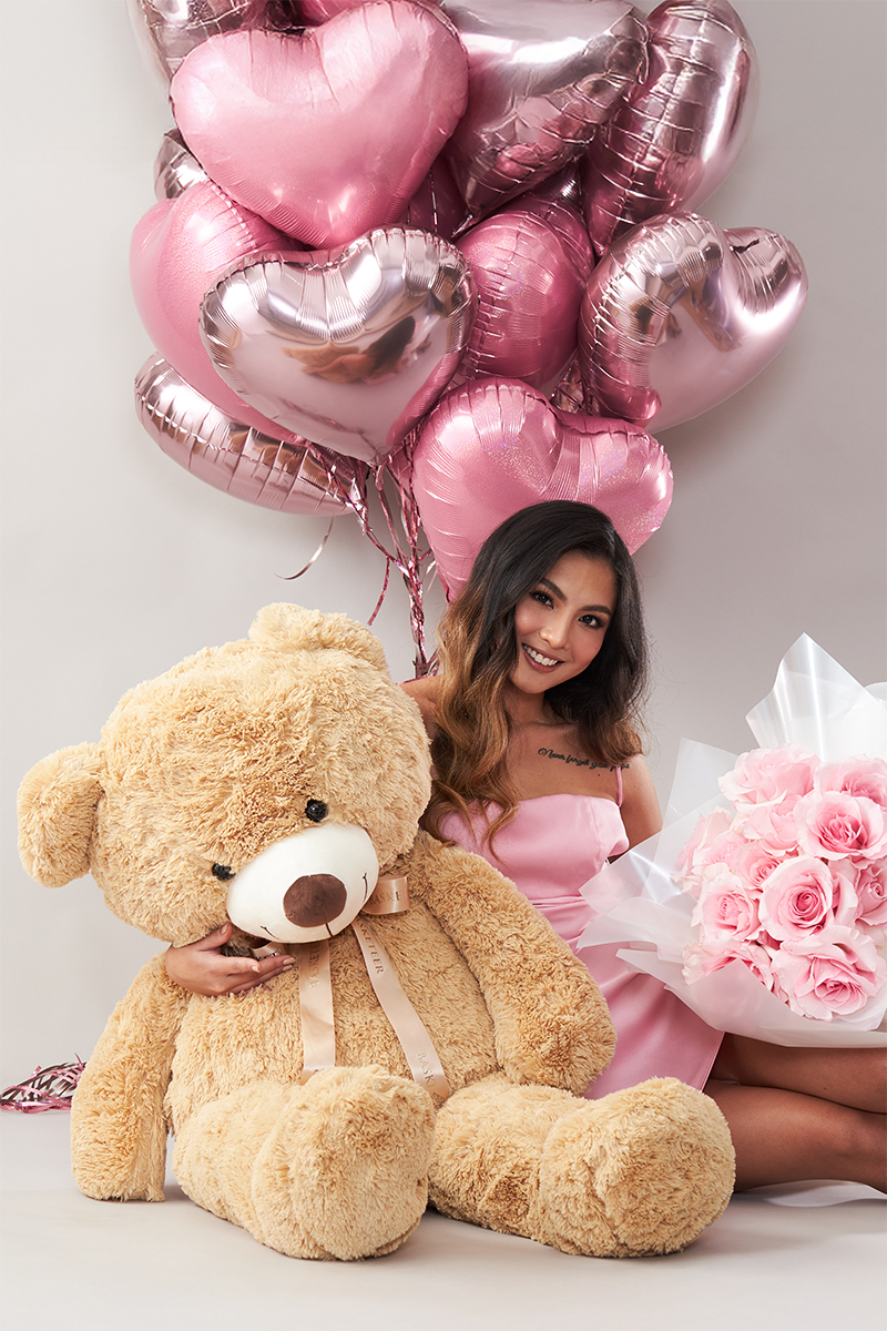 A smiling woman in a pink dress sits holding a large teddy bear, a bouquet of pink roses, and is surrounded by pink heart-shaped balloons.