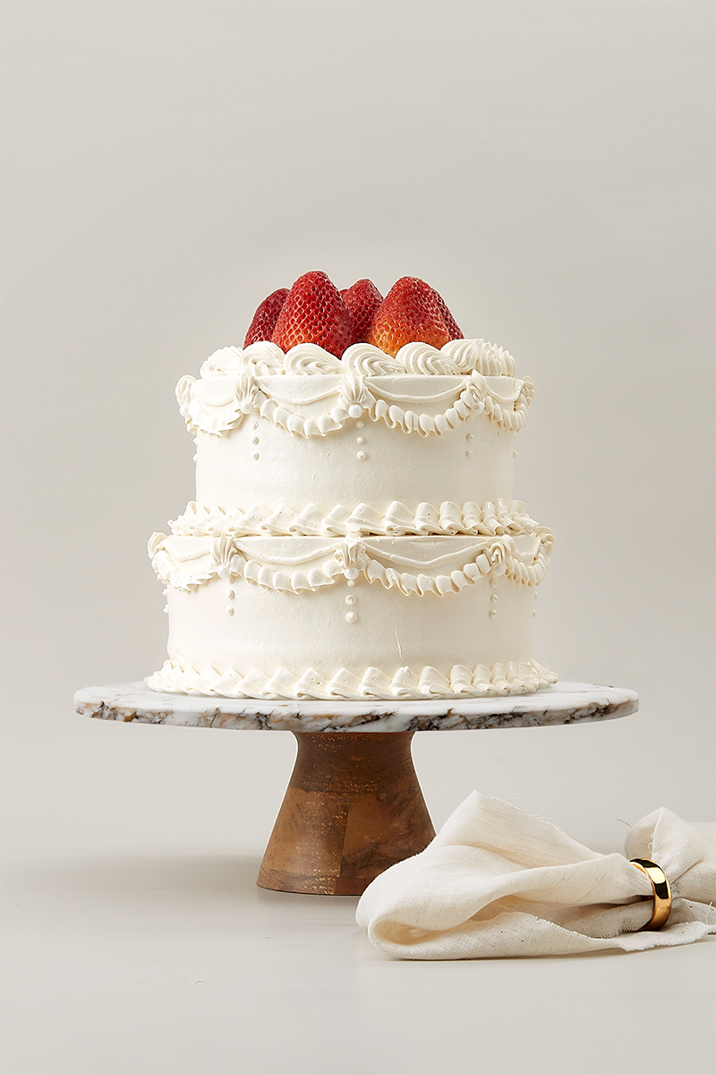 A two-tier vanilla cake with ornate white icing, topped with whole strawberries, sits on a marble cake stand with a wooden base—perfect for your Homepage Select Category. A folded cream-coloured serviette is placed beside the stand.
