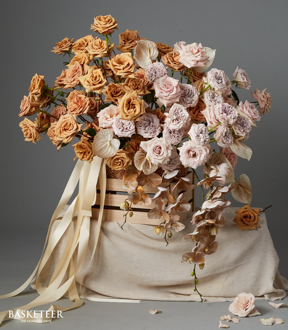 A large, elegant floral arrangement features peach and pale pink roses, light orchids, and pale anthuriums in a round wooden box with long cream ribbons, displayed on a draped beige cloth. “BASKETEER” is in the corner.
