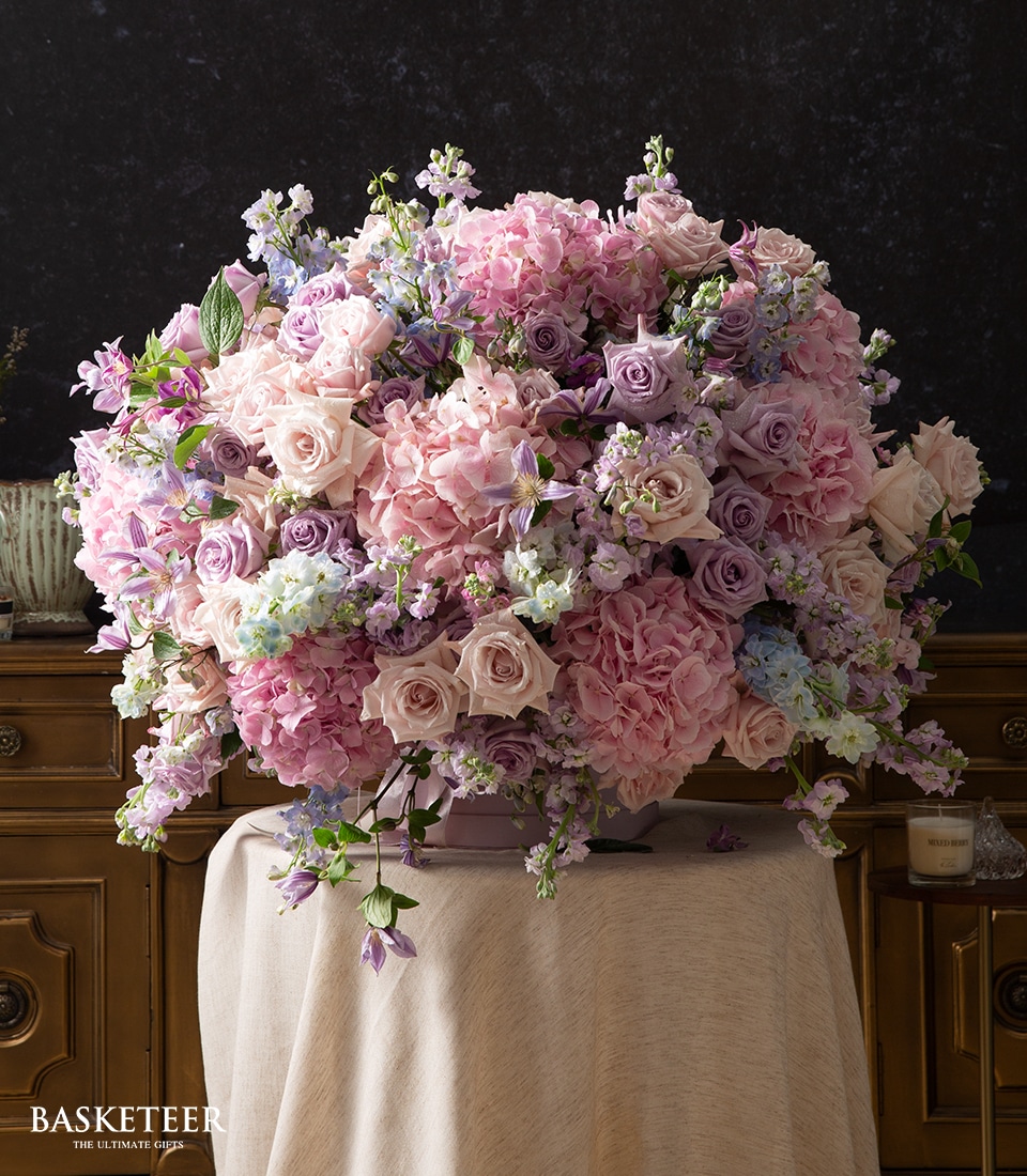 A large, elegant floral arrangement featuring pink and lavender roses, hydrangeas, and other delicate flowers in a round vase, displayed on a draped table against a dark background.