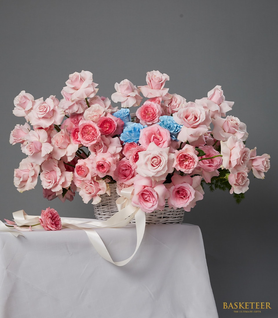 A white wicker basket filled with pink roses and blue flowers sits on a white cloth against a grey background. A white ribbon drapes from the basket. 