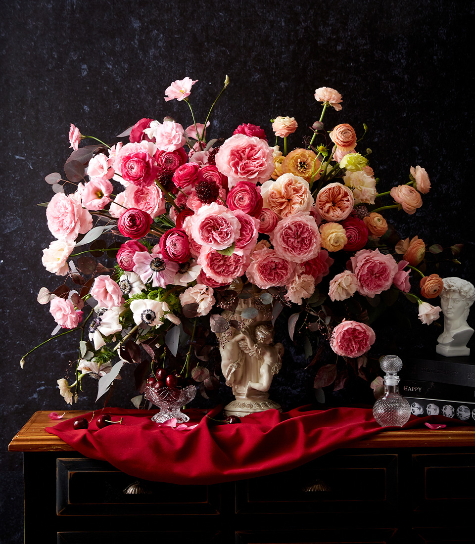 A lush arrangement of pink, red, and peach roses and ranunculus in an ornate vase sits on a dark wooden chest, with crystal decor and a red cloth draped in front, against a dark textured background.