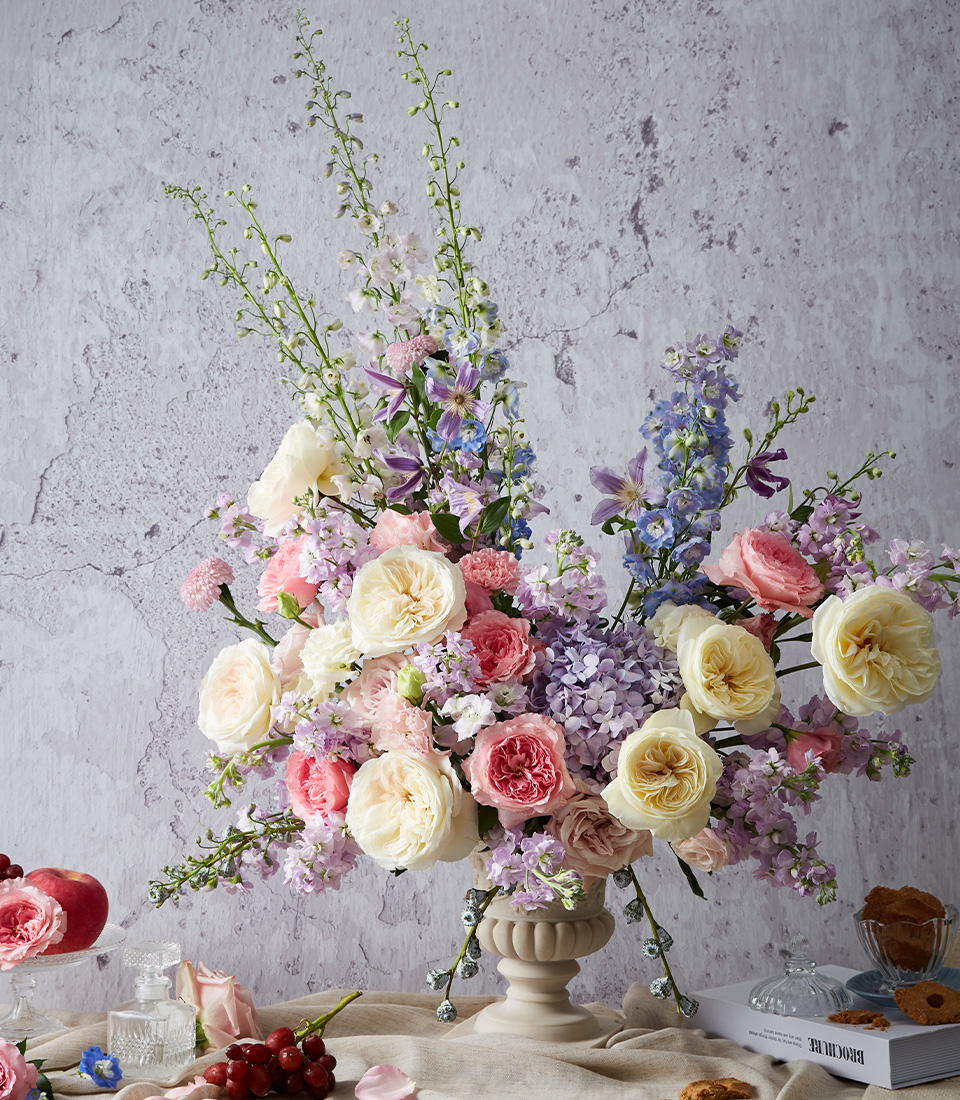 A lush floral arrangement in a cream vase features pastel roses, peonies, and delphiniums in shades of pink, purple, white, and yellow, set on a draped table with fruit and books against a textured light grey wall.