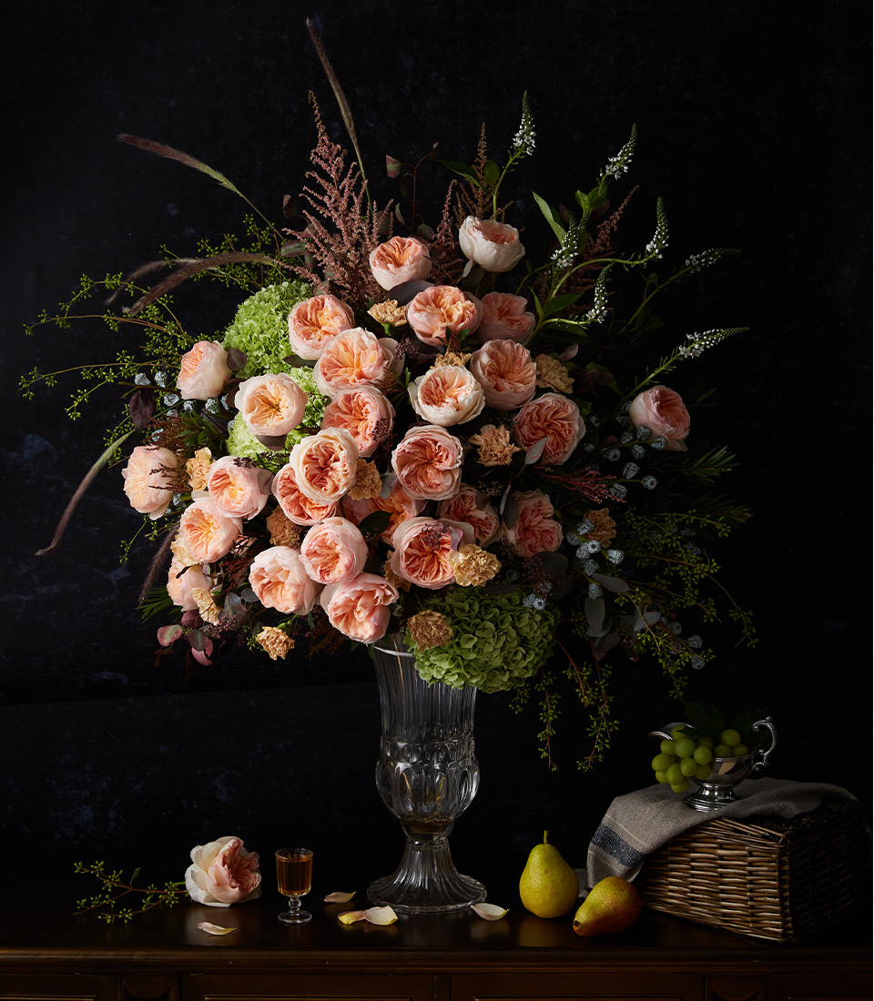 A large bouquet of peach-coloured roses and various greenery in a clear glass vase sits on a wooden table, accompanied by a pear, grapes, a glass of liquor, and a wicker basket with a cloth.