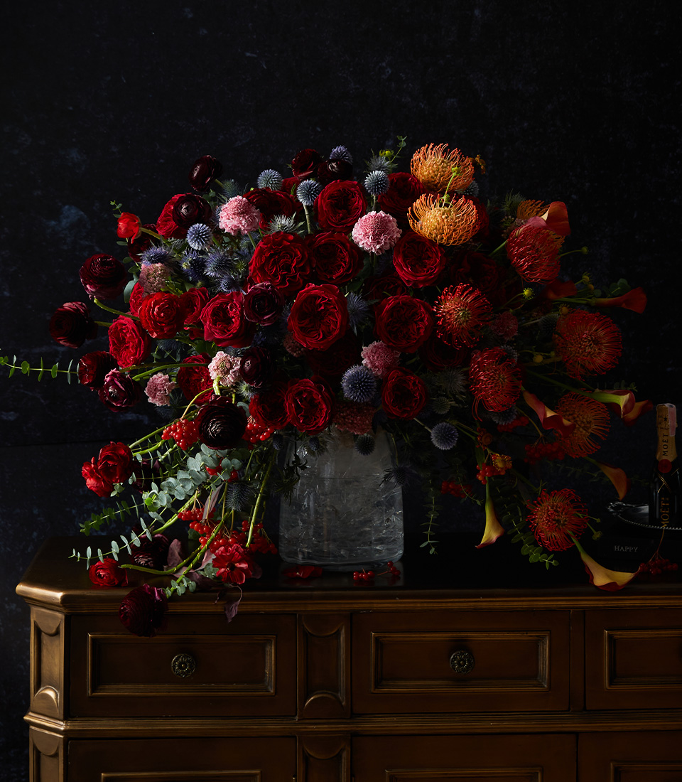 A lush arrangement of deep red, orange, and pink flowers with greenery in a clear glass vase sits on a wooden chest against a dark background.