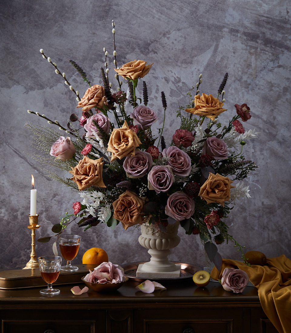 A large floral arrangement in a white vase features mauve, peach, and cream roses, greenery, and pussy willow branches. It sits on a dark table with a candle, fruit, two glasses of liquid, and scattered rose petals.