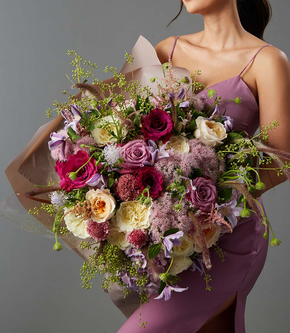 A woman in a mauve dress holds a large, colourful bouquet of roses, carnations, and various greenery against a plain grey background. Her face is not visible.