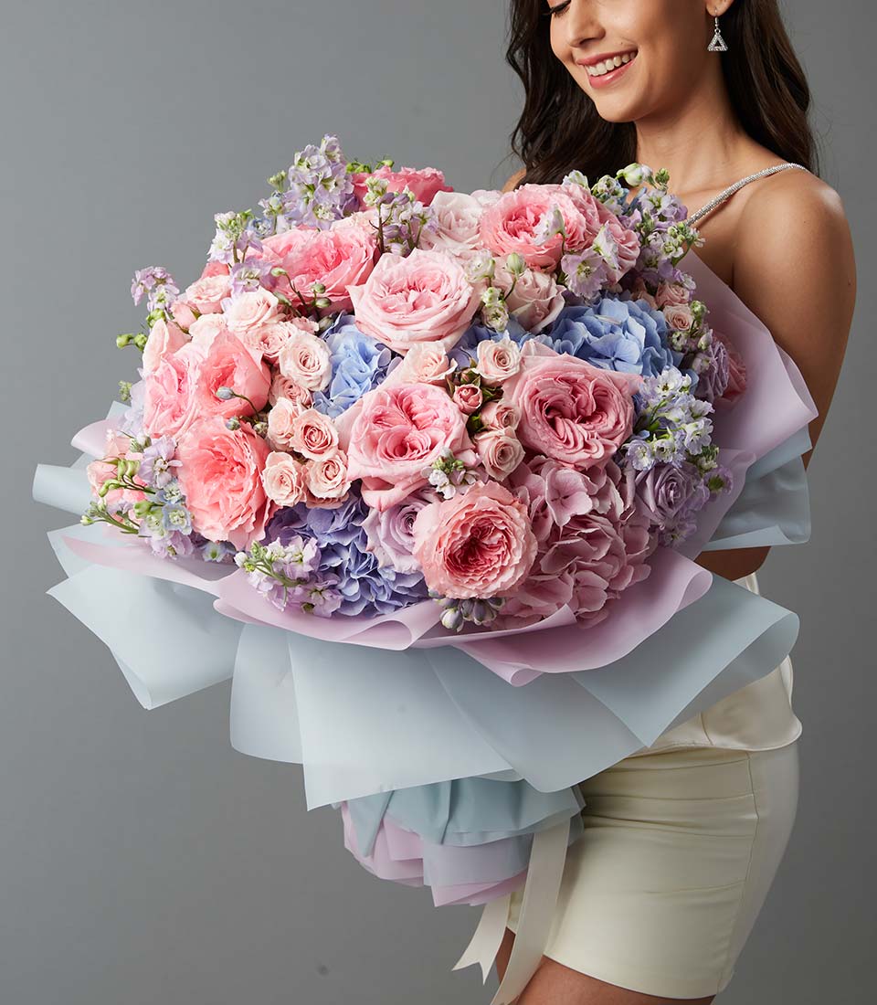A woman in a light dress holds a large bouquet of pink, purple, and blue flowers wrapped in pastel-coloured paper, smiling against a plain grey background.