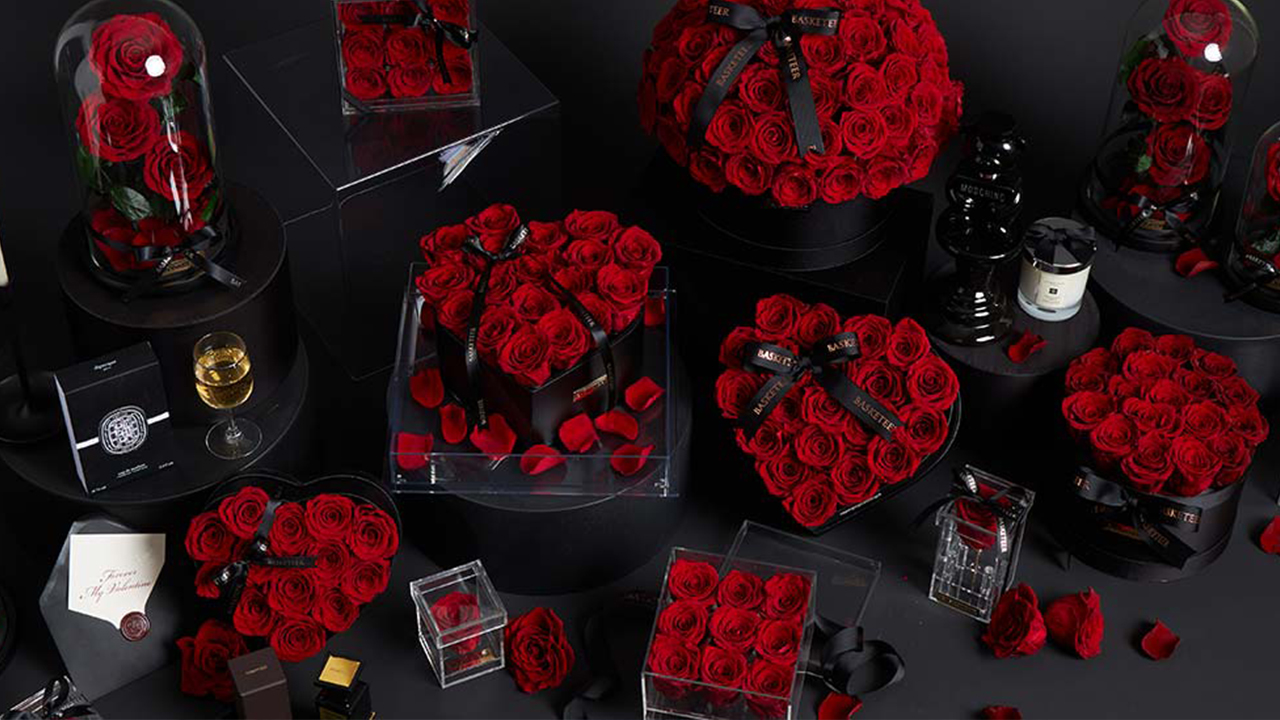A luxurious arrangement of red roses in various shapes—boxes, heart, domes—displayed with black gift boxes, glass containers, candles, and scattered rose petals on a dark surface.